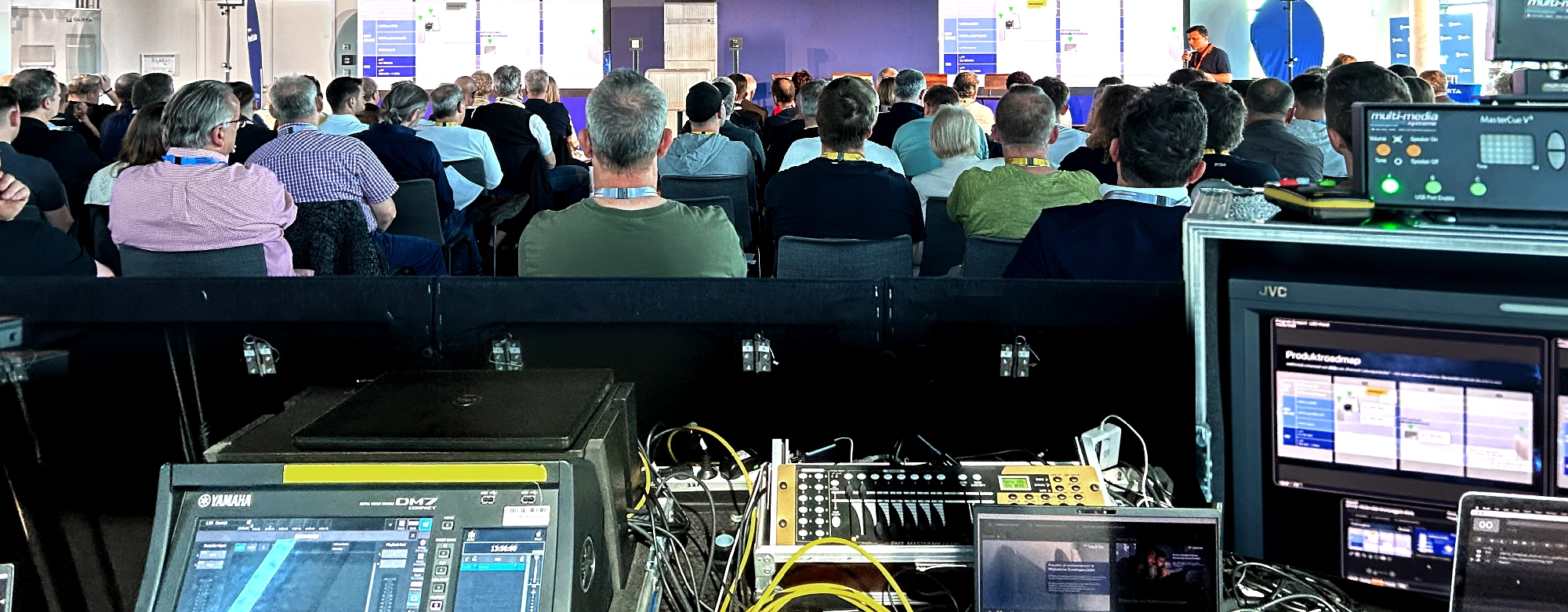 Im Vordergrund ein Aufbau der Veranstaltungstechnik. Im Hintergrund das Publikum eines Business Events, welches auf die Bühne schaut.